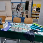 Stoisko WSzS podczas targów. Na pierwszym planie jest stolik przykryty zielonym płótnem. Na nim leżą materiały promocyjne szpitala: teczki, kwartalnik Twój Lekarz, smycze, breloczki oraz inne.Przy stoliku 2 krzesła. W tle roll-up szpitala.