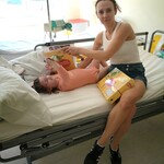 Na łóżku szpitalnym leży mała dziewczynka. Obok siedzi mama trzymając upominek z okazji Dnia Dziecka.