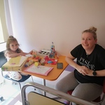 Dziewczynka oraz mama siedzą na krzesełkach przy stoliku, na którym znajdują się kolorowanka oraz otrzymany upominek z okazji Dnia Dziecka.