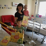 Mama stoi przy łózku szpitalnym. Na rękach trzyma małe dziecko. Obok nich stoi chłopczyk, który trzyma prezent z okazji Dnia Dziecka.